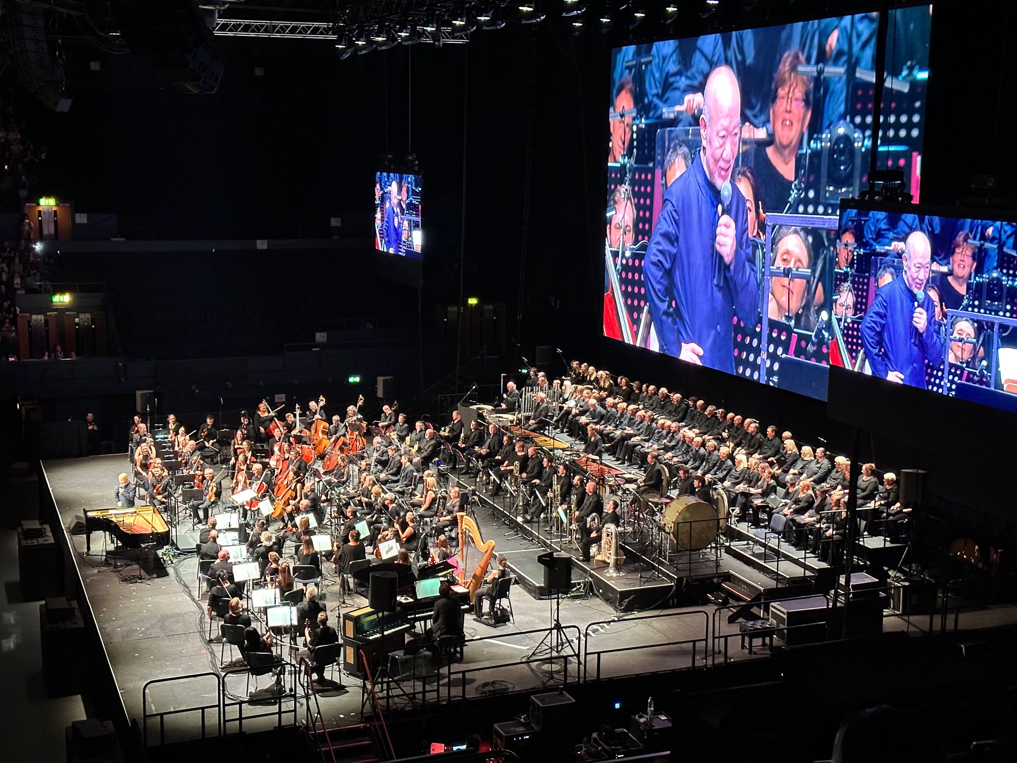 Joe Hisaishi addressing the audience with a mic in front of the symphonic orchestra, his image being live broadcast on the big screens in the background as well 